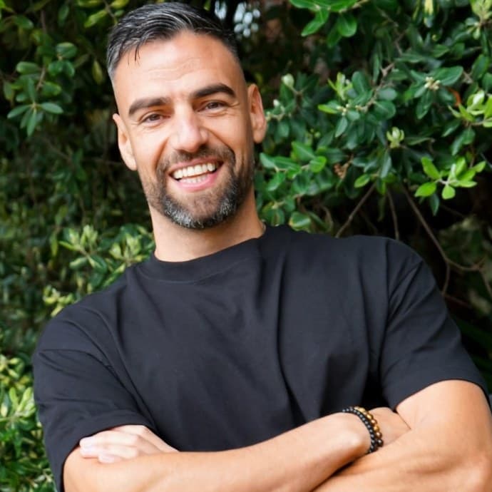 Mike Henderson grinning, arms folded, in front of green foliage